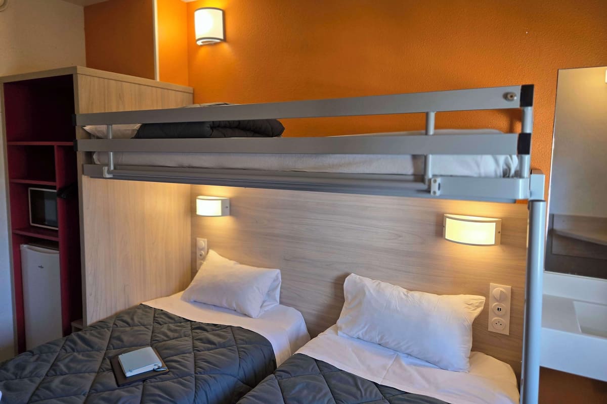 A bunk bed room with two beds, each with a pillow and a quilted blanket. There are wall-mounted lights above the beds and a small table with a book.