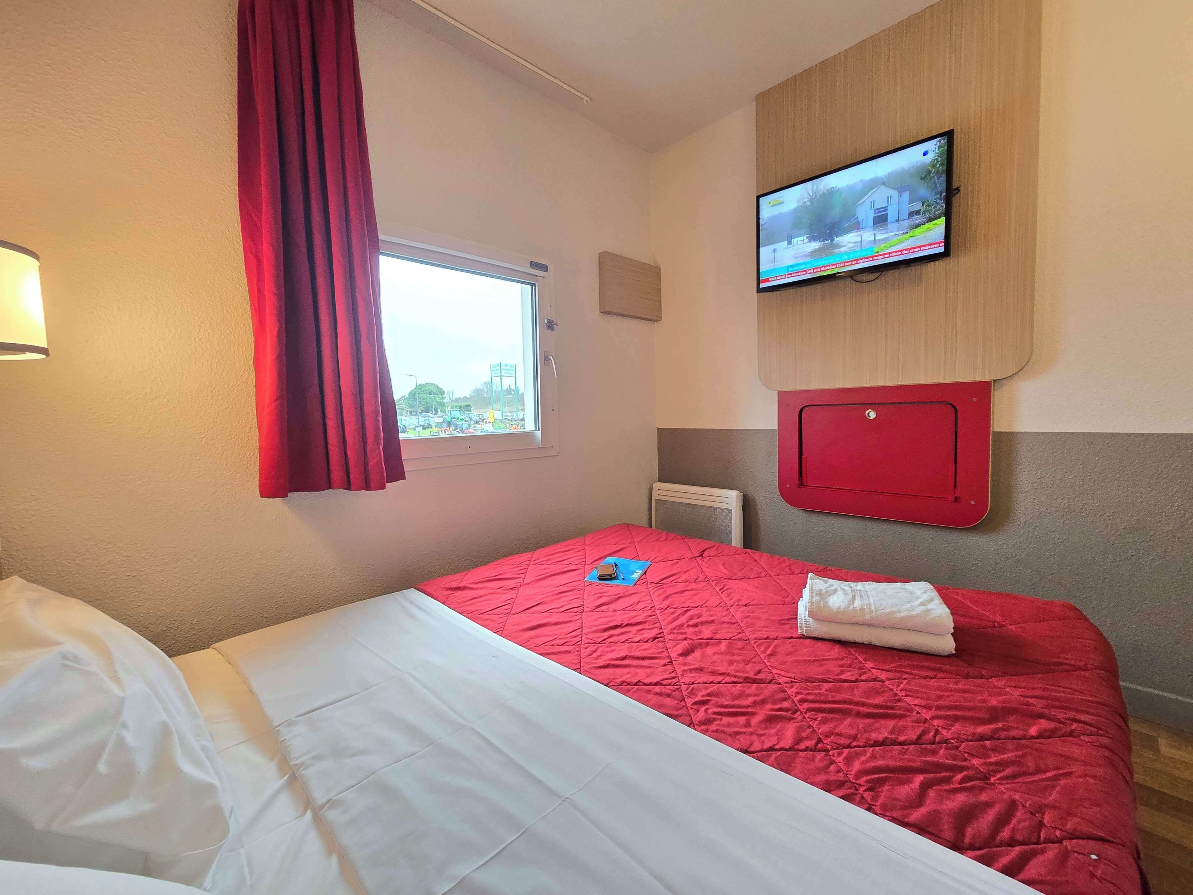 Habitación de hotel con cama, televisor montado en la pared, ventana con cortinas y radiador.