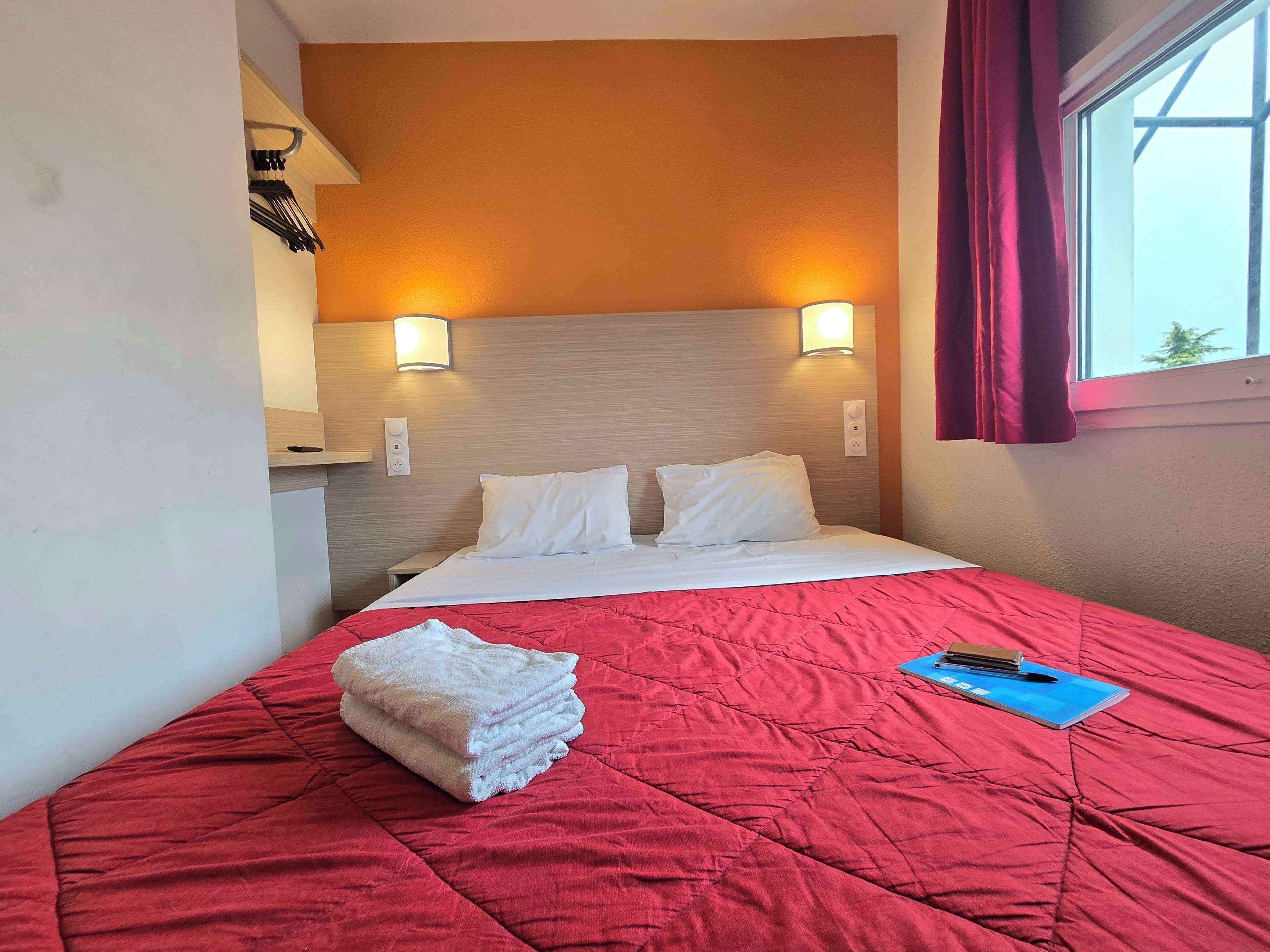 Une chambre d’hôtel avec un lit, une couverture matelassée rouge, une pile de serviettes blanches et un carnet bleu posé sur le lit.