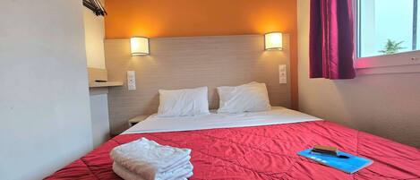 A hotel room with a bed, a red quilted bedspread, a stack of white towels, and a blue notebook on the bed.