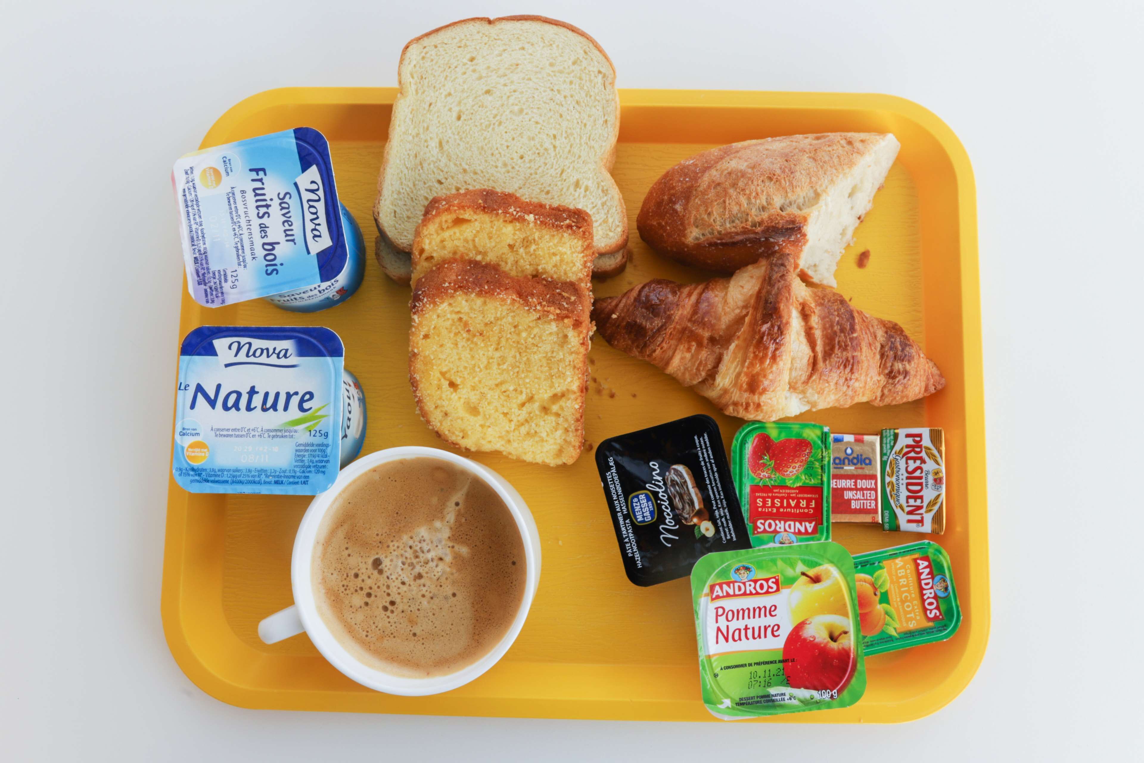 Una bandeja con rebanadas de pan, un croissant, una taza de café y varios alimentos, incluyendo yogur y fruta.