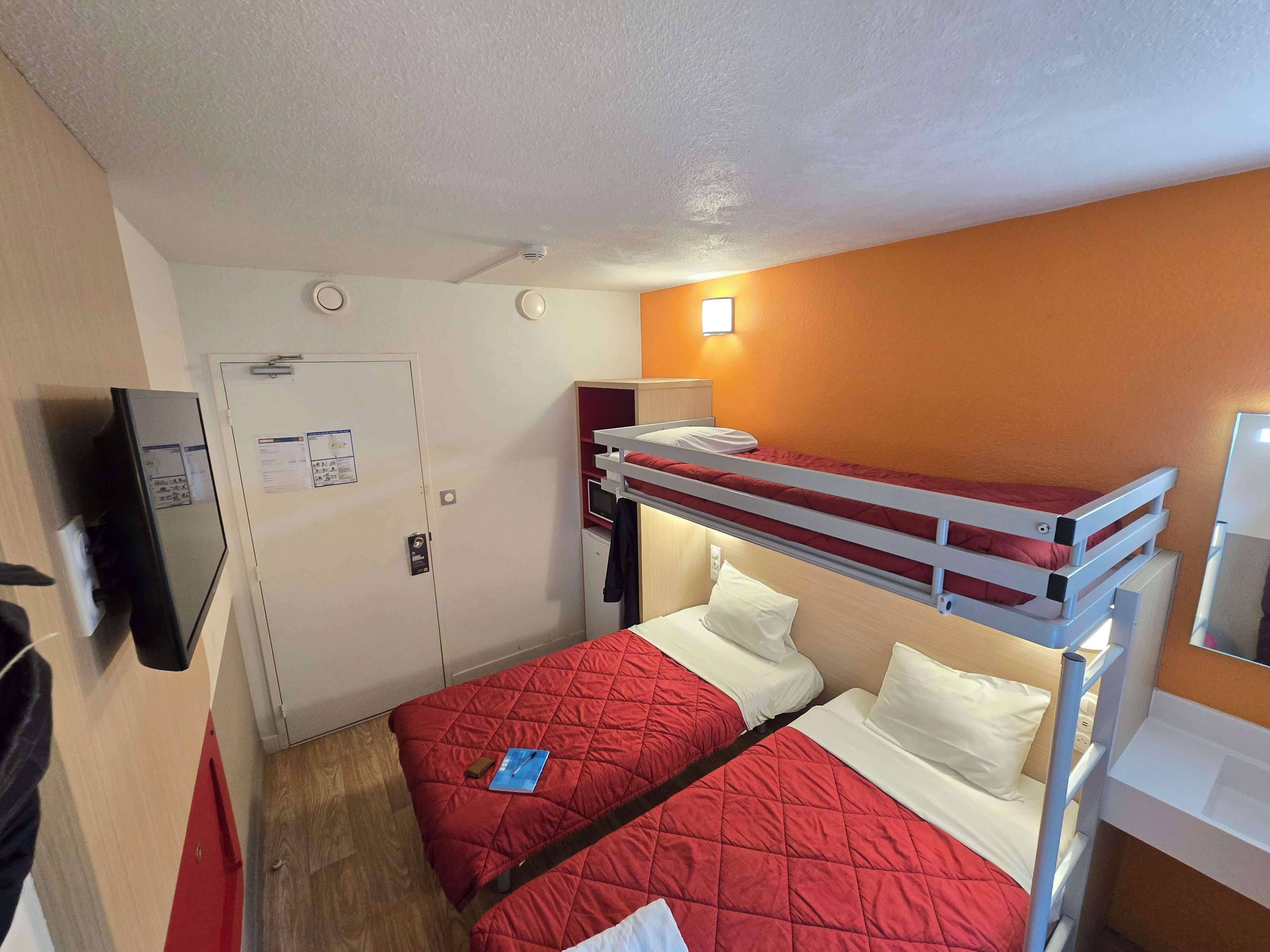 A bunk bed room with a red quilted bedspread, a white pillow, a small blue book, and a white door with a bulletin board.
