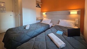A hotel room with two beds, each with a grey quilted bedspread and white pillows. There are two bedside tables, one with a book and a laptop on it. The room has a wooden headboard, a wall-mounted light, and a door with a handle.