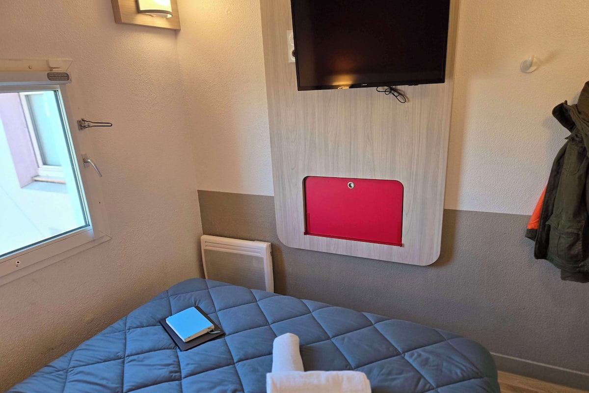 A room with a bed, a window, a wall-mounted TV, and a red panel.