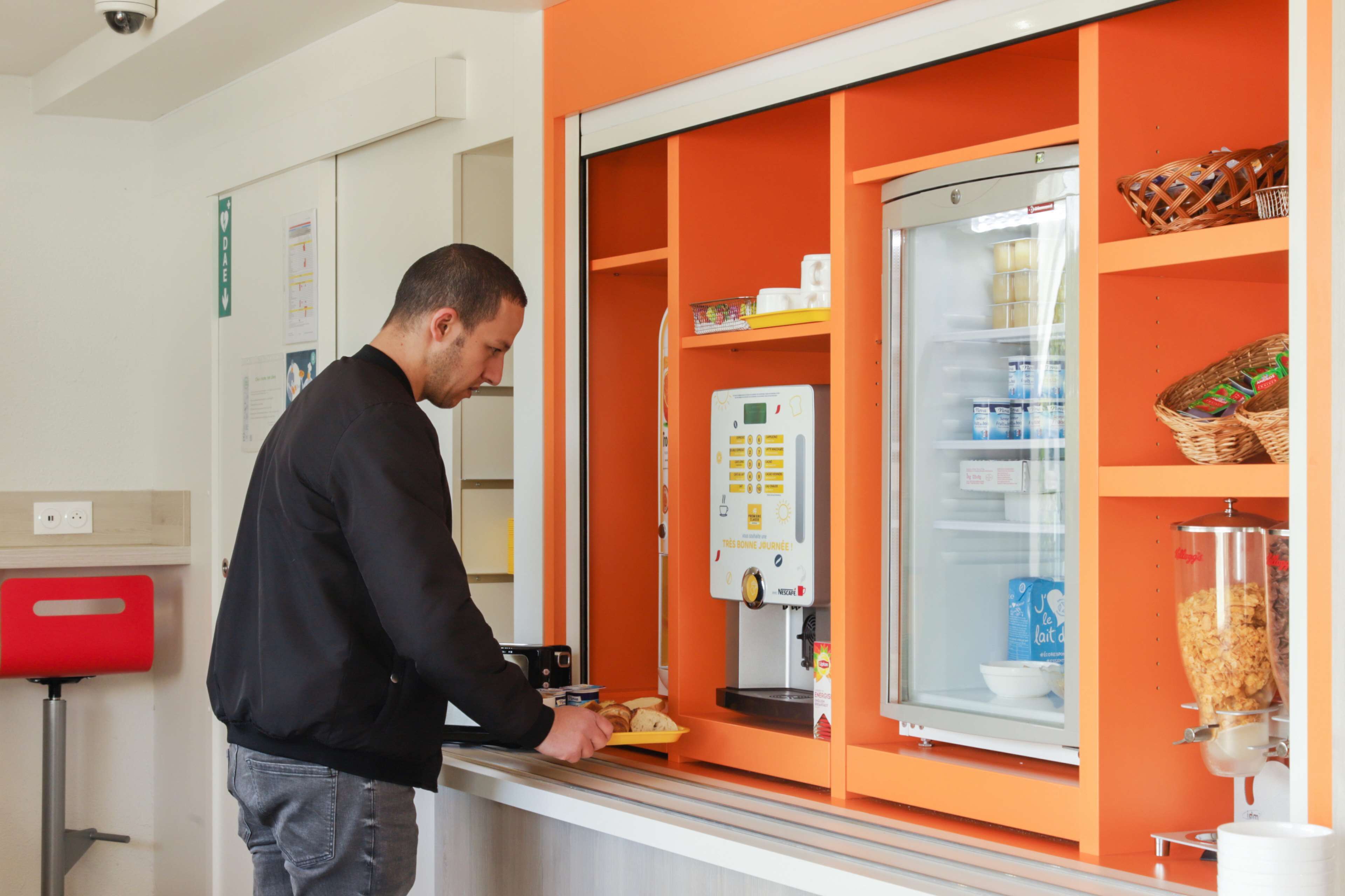A man at a self-service breakfast station with a refrigerator stocked with beverages and a coffee machine.