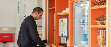 A man at a self-service breakfast station with a refrigerator stocked with beverages and a coffee machine.