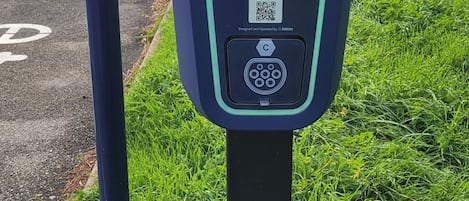 Una estación de carga para vehículos eléctricos con la marca 'etotem', ubicada en un área con césped, una valla y edificios industriales al fondo.