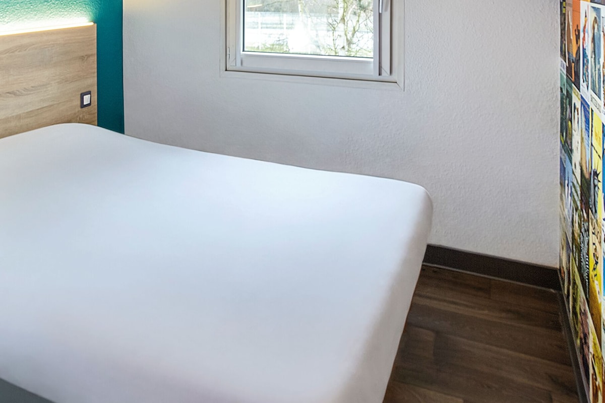 A room with a white bed, a wooden headboard, a small window, and a wall with a light switch.