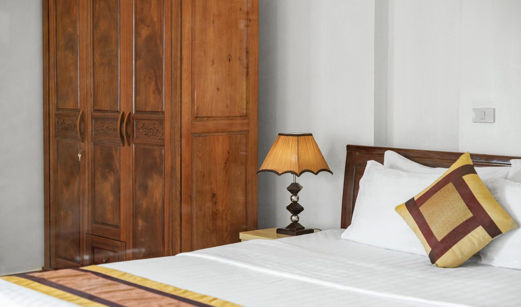 A hotel room with a large wooden wardrobe, a bedside lamp, a bed with white bedding, and a decorative pillow.
