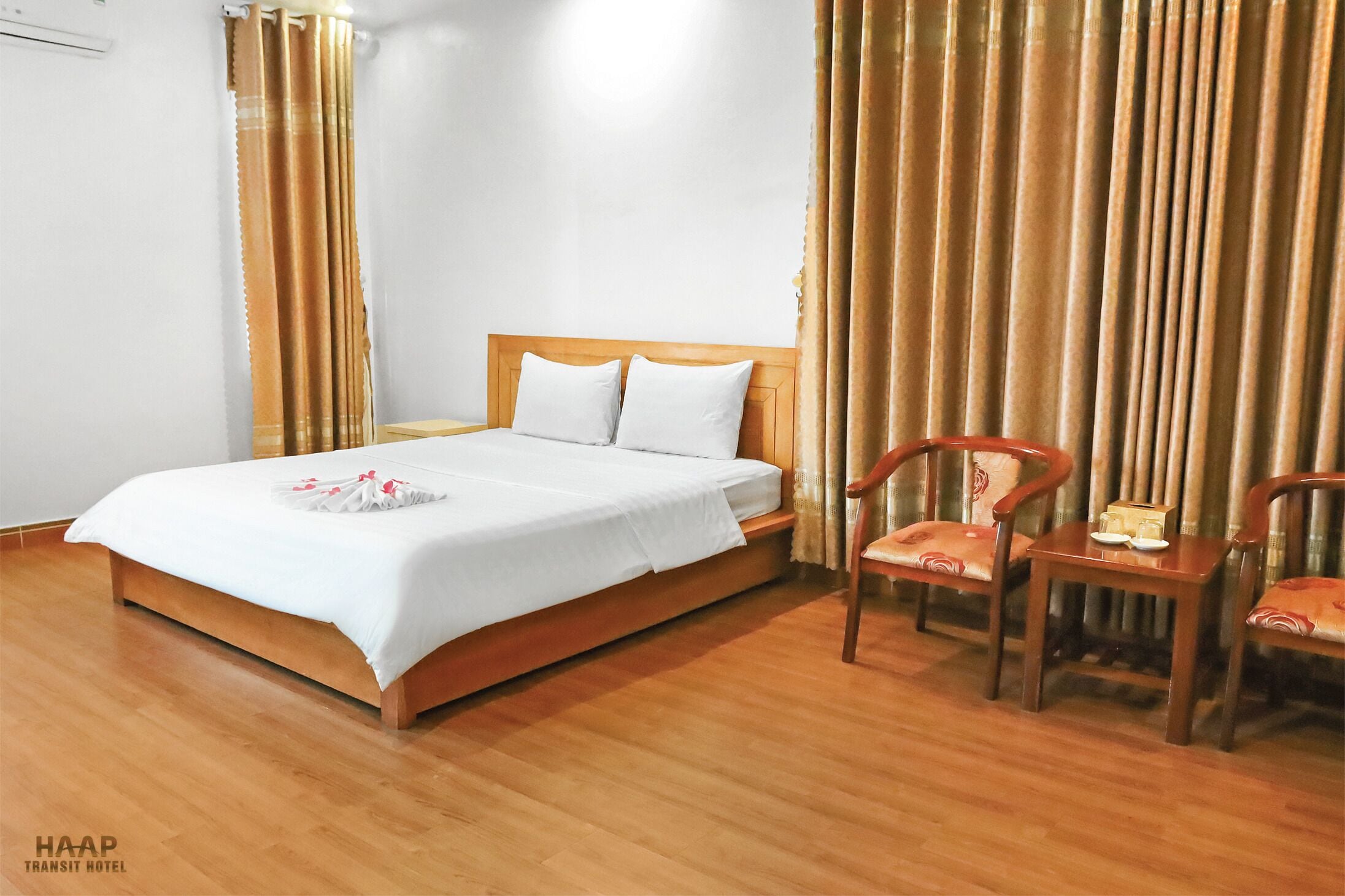 Une chambre d’hôtel avec un lit en bois, du linge de lit blanc, deux chaises en bois et une petite table sur laquelle se trouvent une boîte et une assiette.