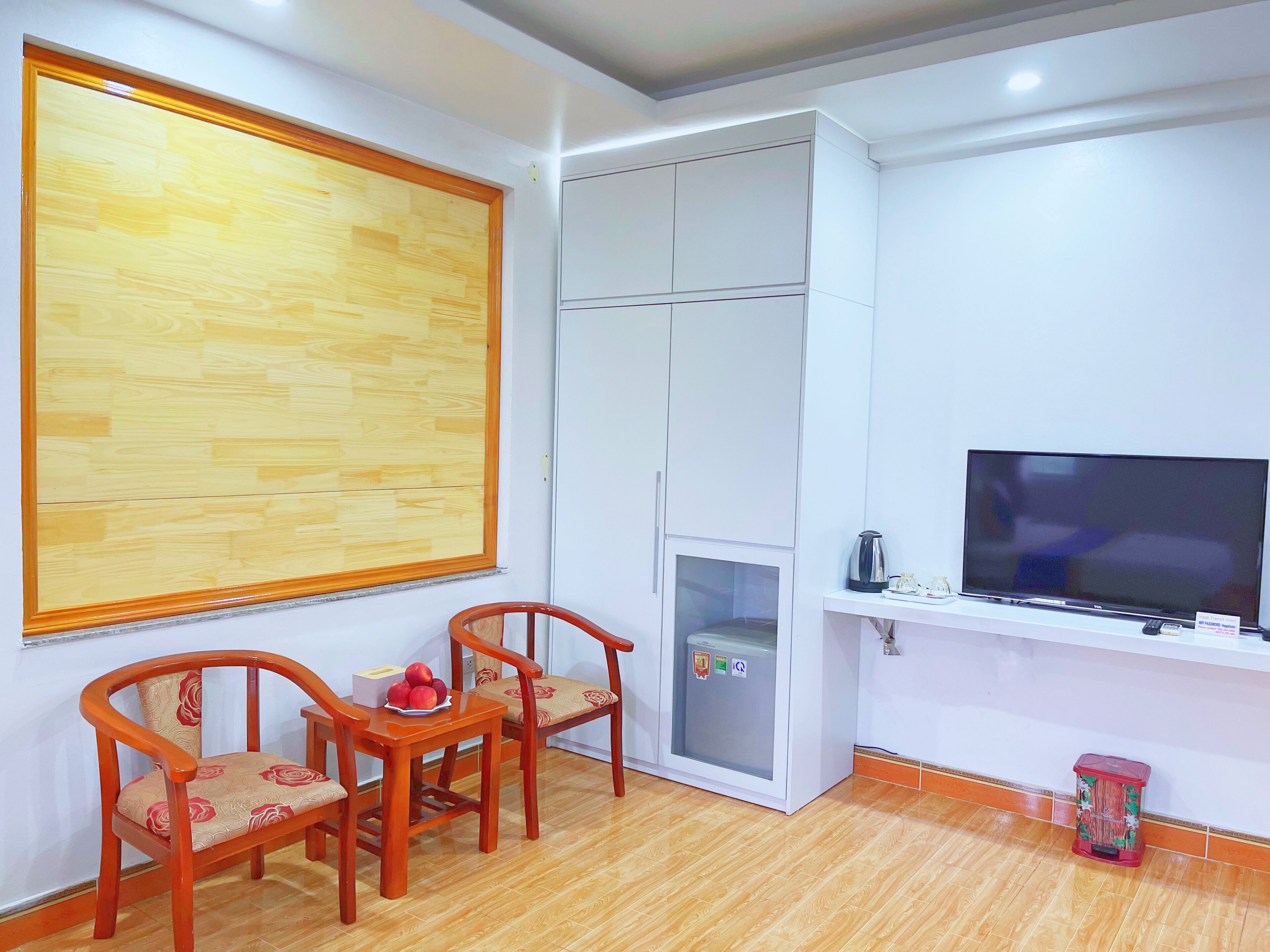 A room with a wooden floor, a white wardrobe, a television on a stand, and two wooden chairs with cushions.