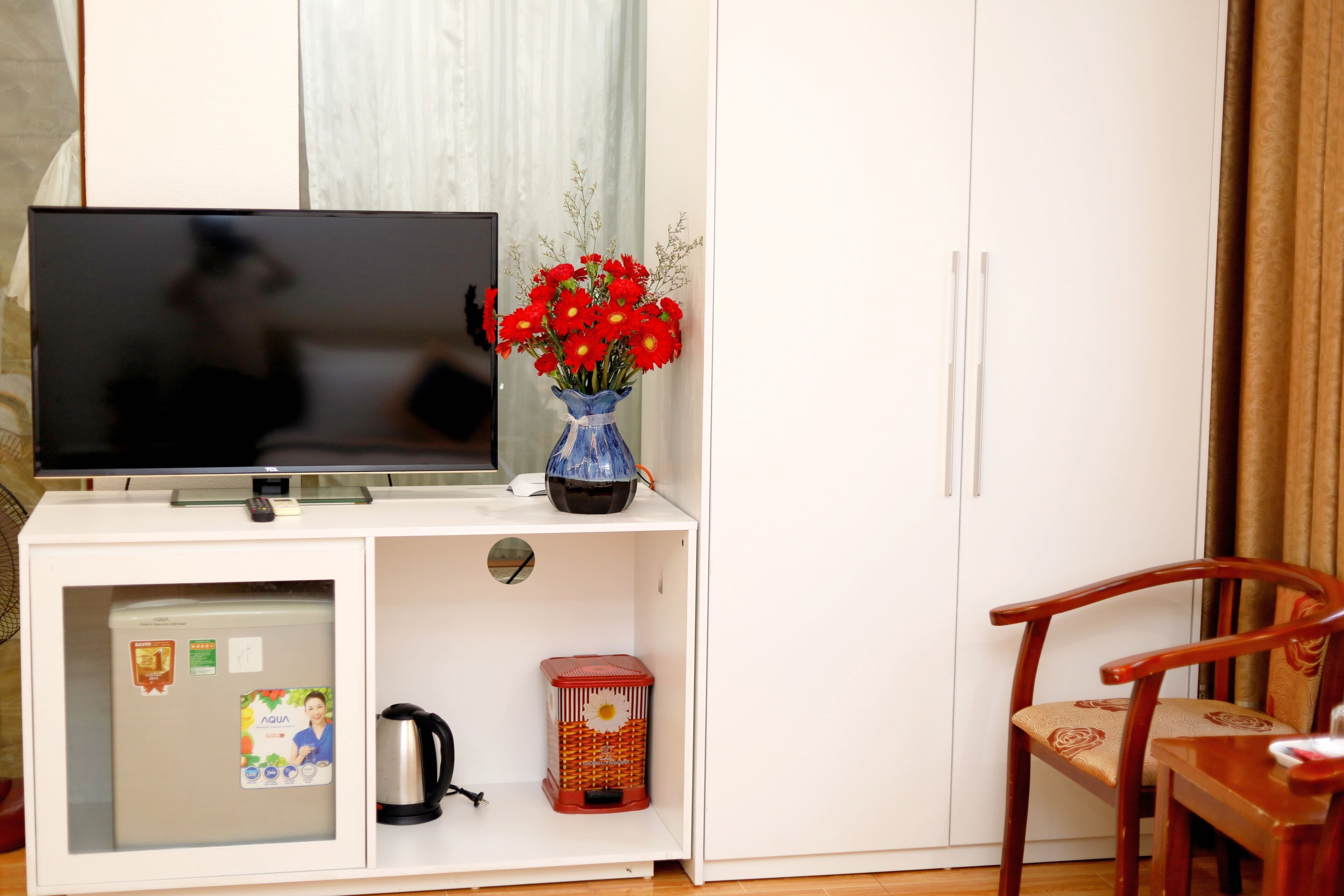 Una habitación con una televisión sobre un mueble blanco, un jarrón con flores rojas, un farolillo rojo y una silla de madera.