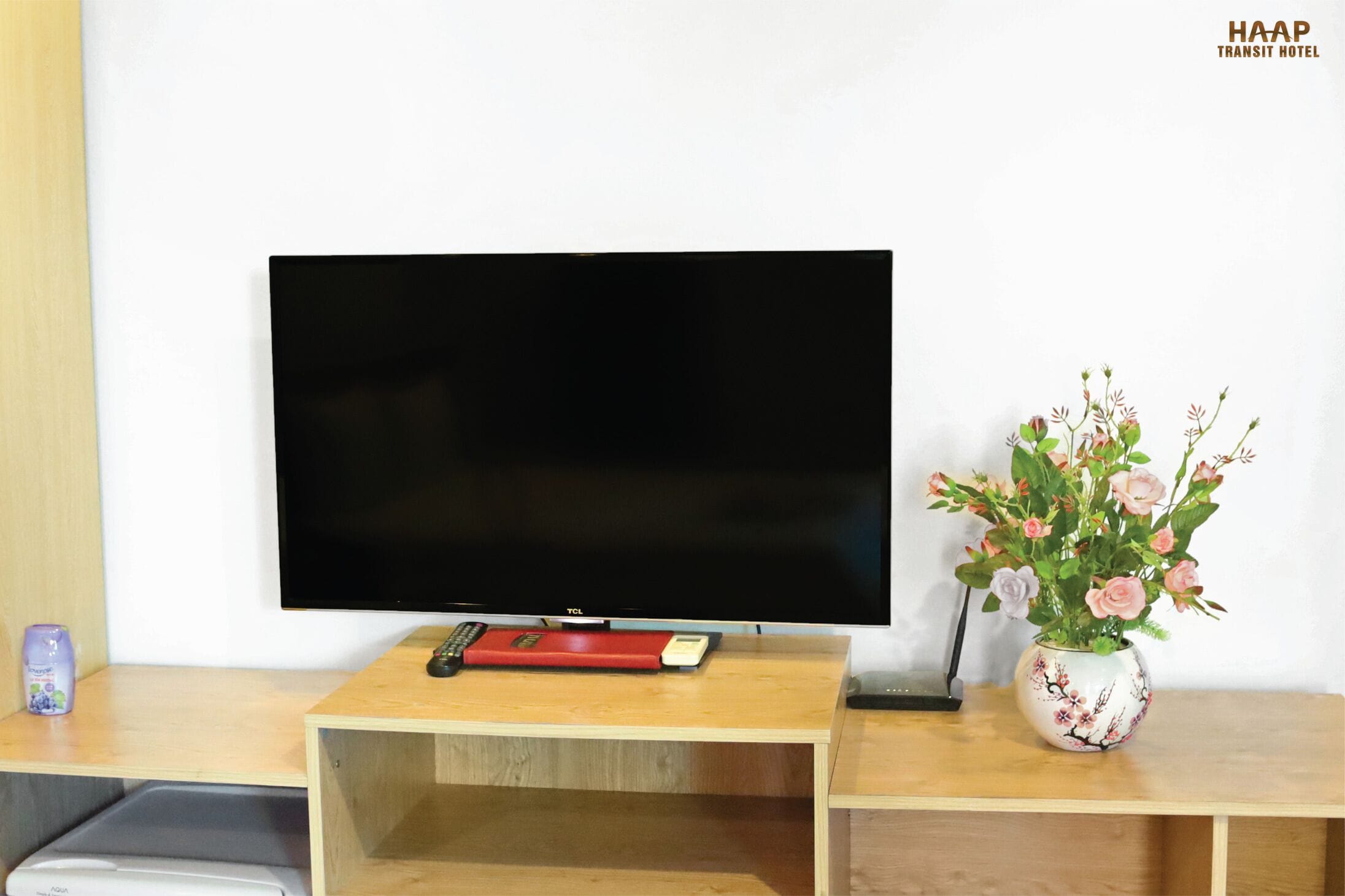 Een flatscreen televisie op een houten standaard, samen met een afstandsbediening en een decoratieve vaas met bloemen.