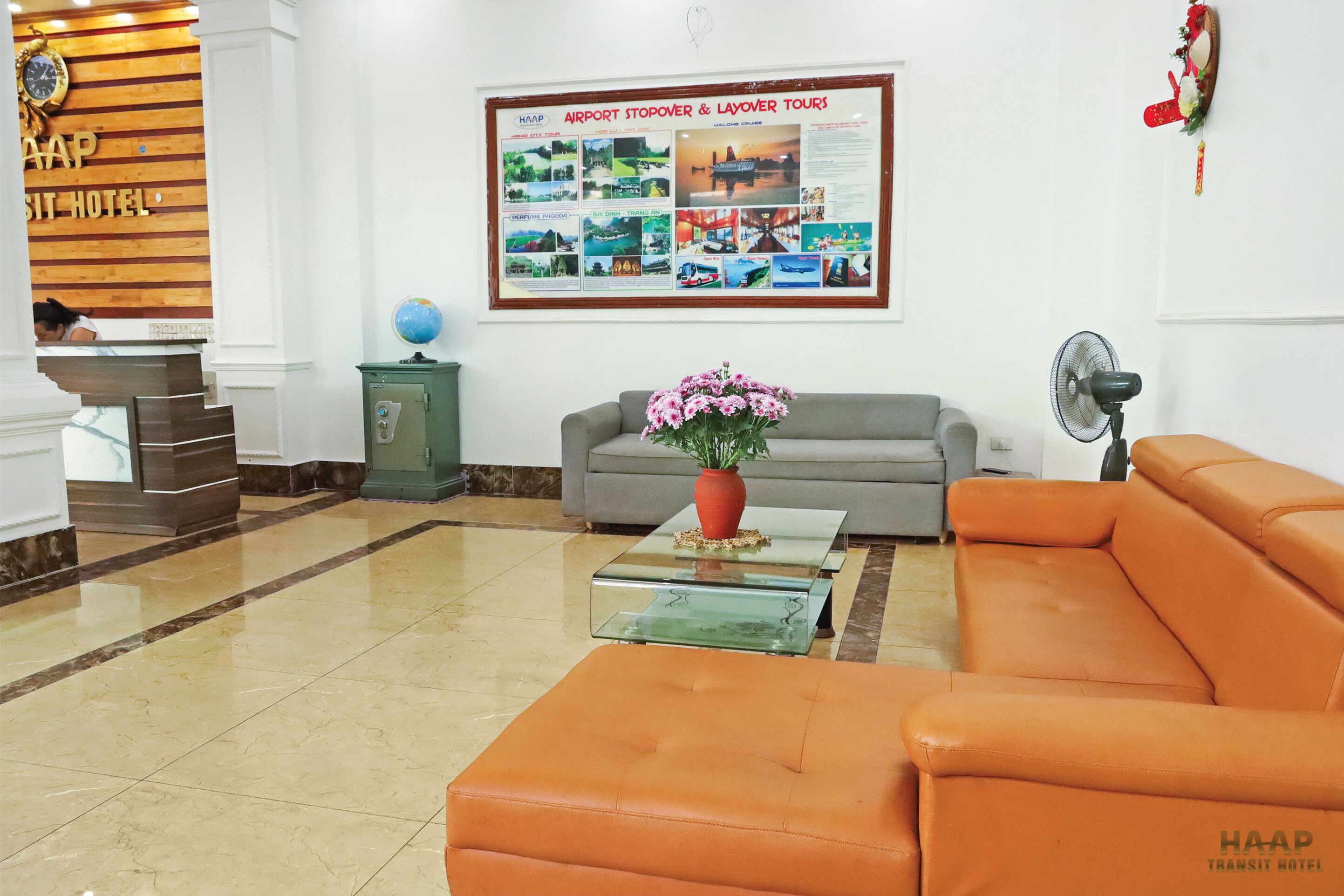 A hotel lobby with a reception desk, a sofa, a coffee table with a vase of flowers, and a poster advertising tours.