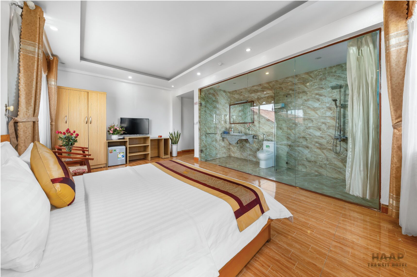 A modern hotel room with a large bed, a TV, a desk, a wardrobe, a bathroom with a glass shower, and a view of the outside.