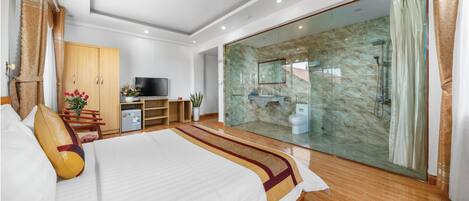 A modern hotel room with a large bed, a TV, a desk, a wardrobe, a bathroom with a glass shower, and a view of the outside.