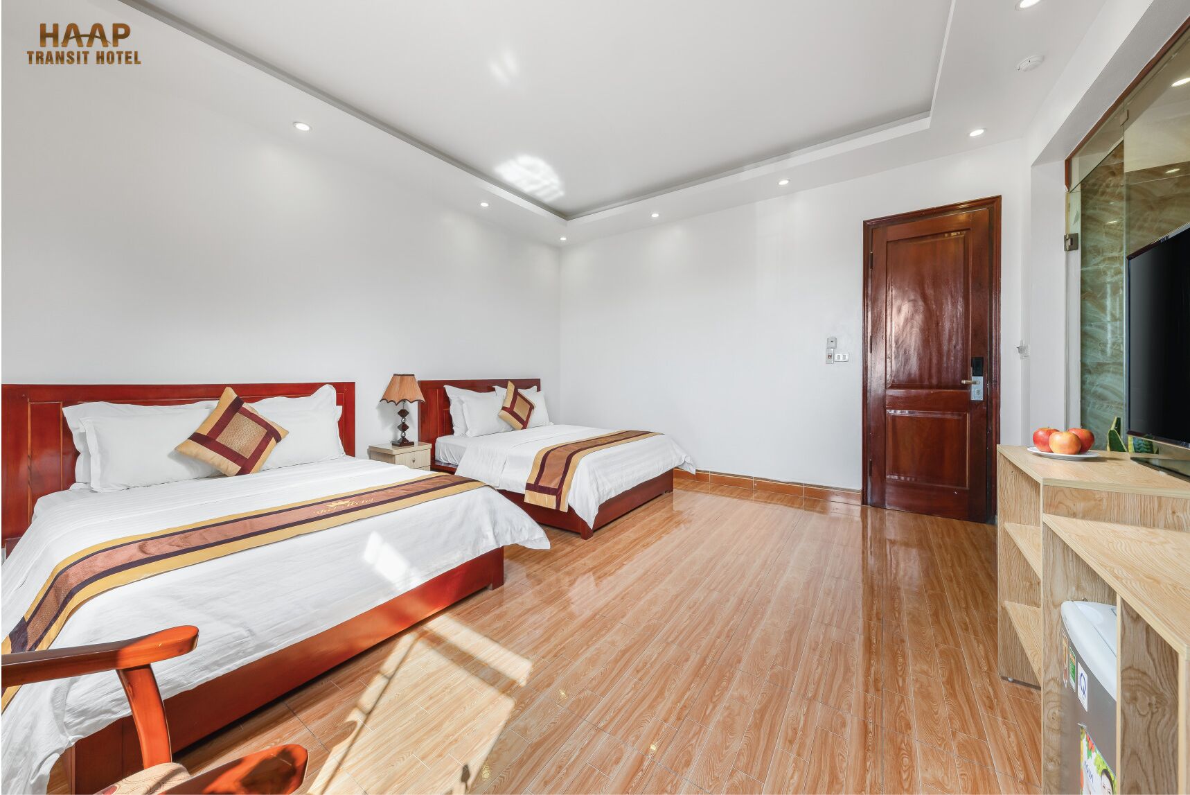 Une chambre d’hôtel avec deux lits, un plancher en bois, une porte en bois et un téléviseur posé sur une commode en bois.