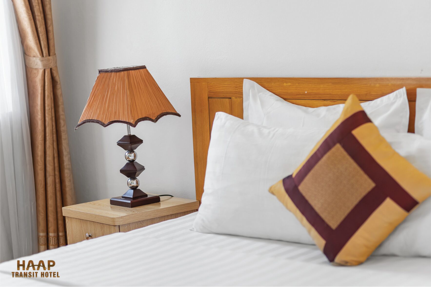 Habitación de hotel con mesita de noche de madera, lámpara, cama con ropa blanca y un cojín decorativo.