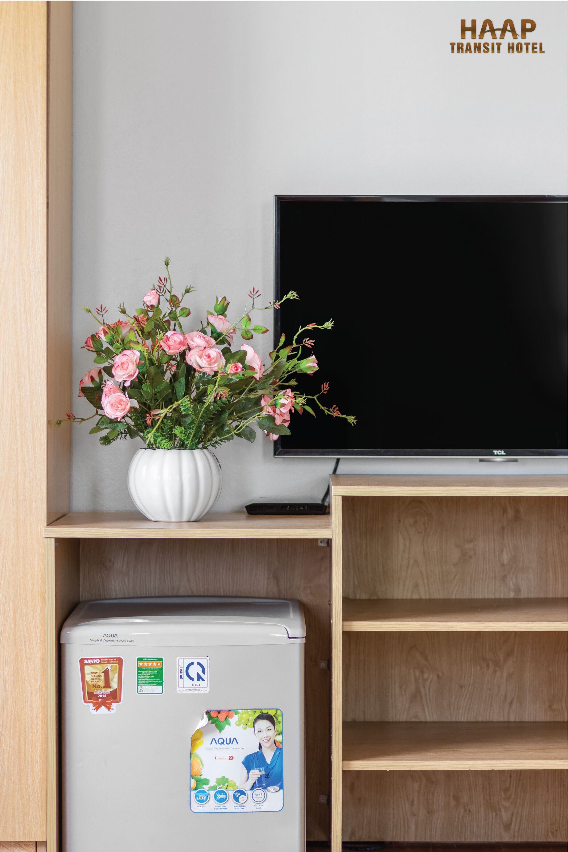 A hotel room with a flat-screen TV, a white vase with pink flowers, and a refrigerator with various stickers.