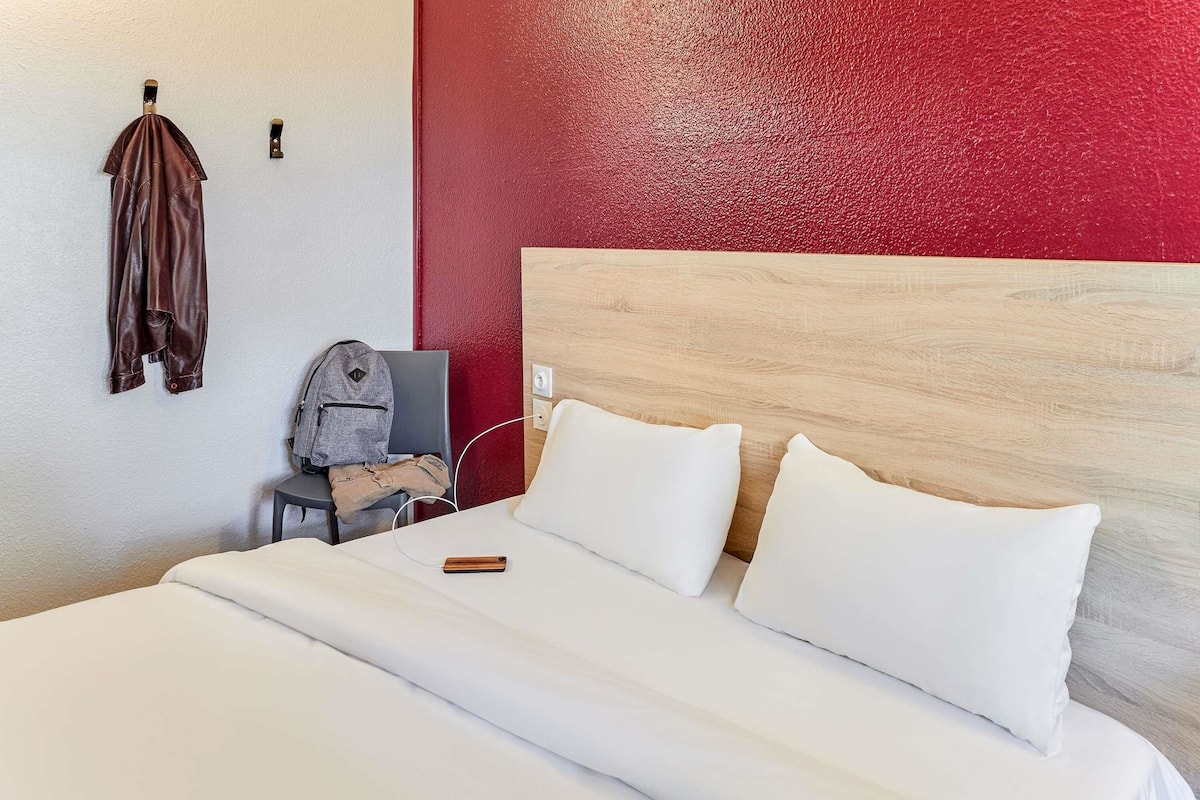 Habitación de hotel con cama, almohadas blancas, cabecero de madera, pared de acento roja, perchero y una mochila en una silla.