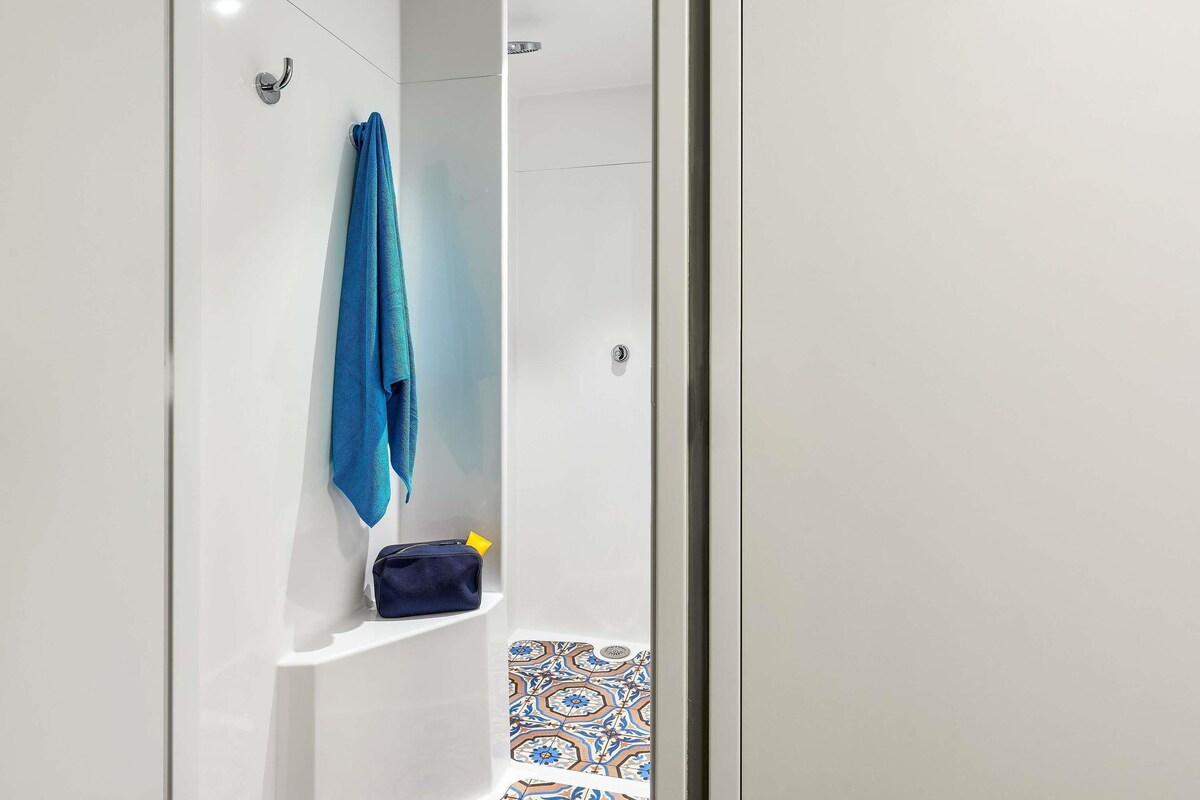 Un baño con pared blanca, una toalla azul colgada en un gancho y un estante con un bolso y un objeto amarillo.