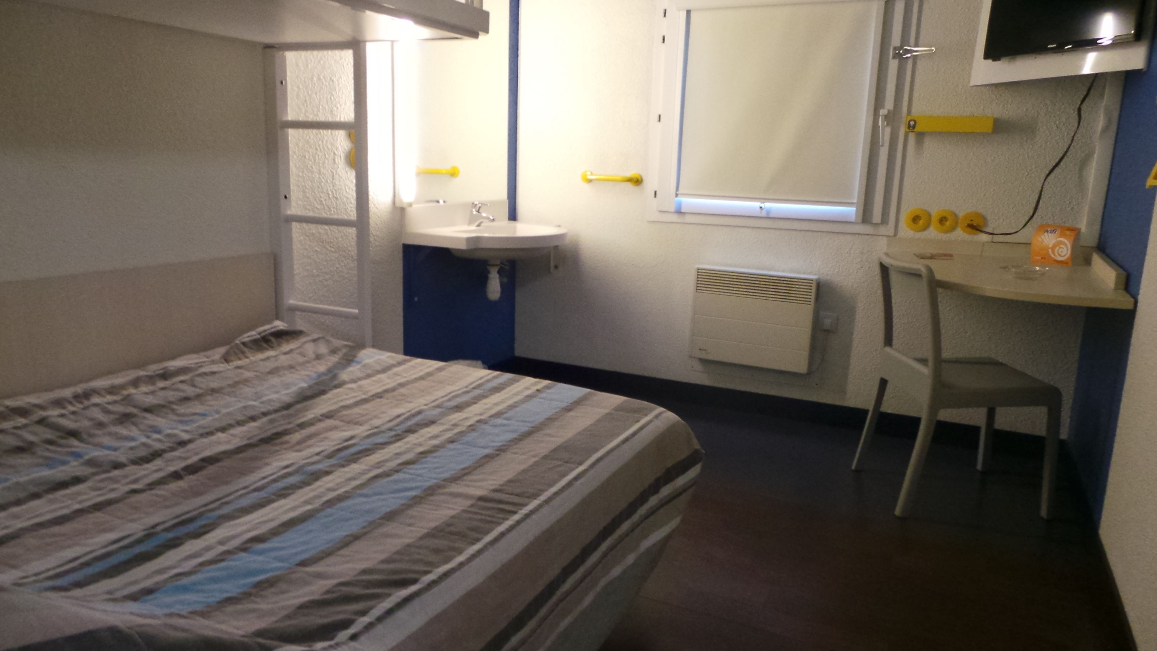 Ein Hotelzimmer mit einem Bett, einem Schreibtisch, einem Stuhl, einem Waschbecken und einem an der Wand befestigten Fernseher.