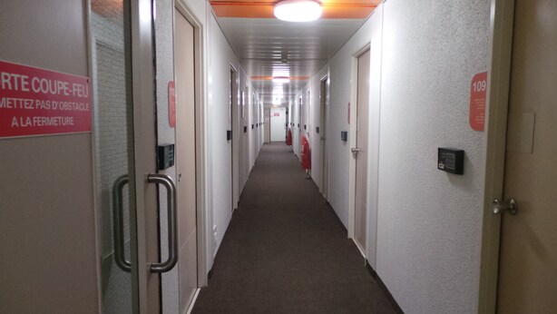 Ein langer, schmaler Hotelkorridor mit Türen an beiden Seiten, einem Feuerlöscher und einem Schild an der linken Wand.