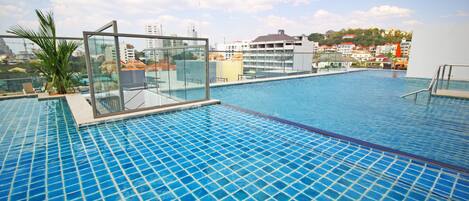 Une piscine à débordement située sur le toit, entourée de parois en verre, avec une vue sur les bâtiments de la ville et un ciel dégagé.