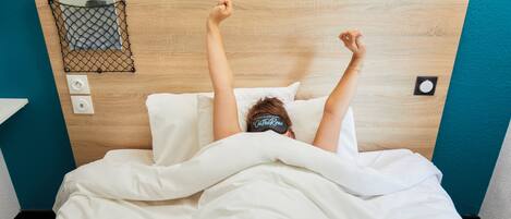 Una persona acostada en la cama con los brazos levantados, un cartel con la palabra "ROADITUDE" en la cabecera y un enchufe en la pared.
