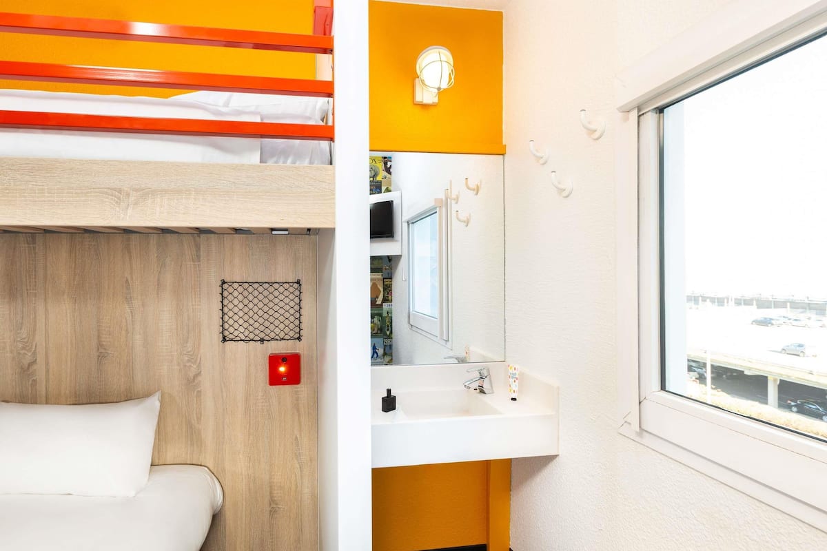 A bunk bed with a wooden headboard, a white sink with a mirror above it, and a window with a view of a parking lot.