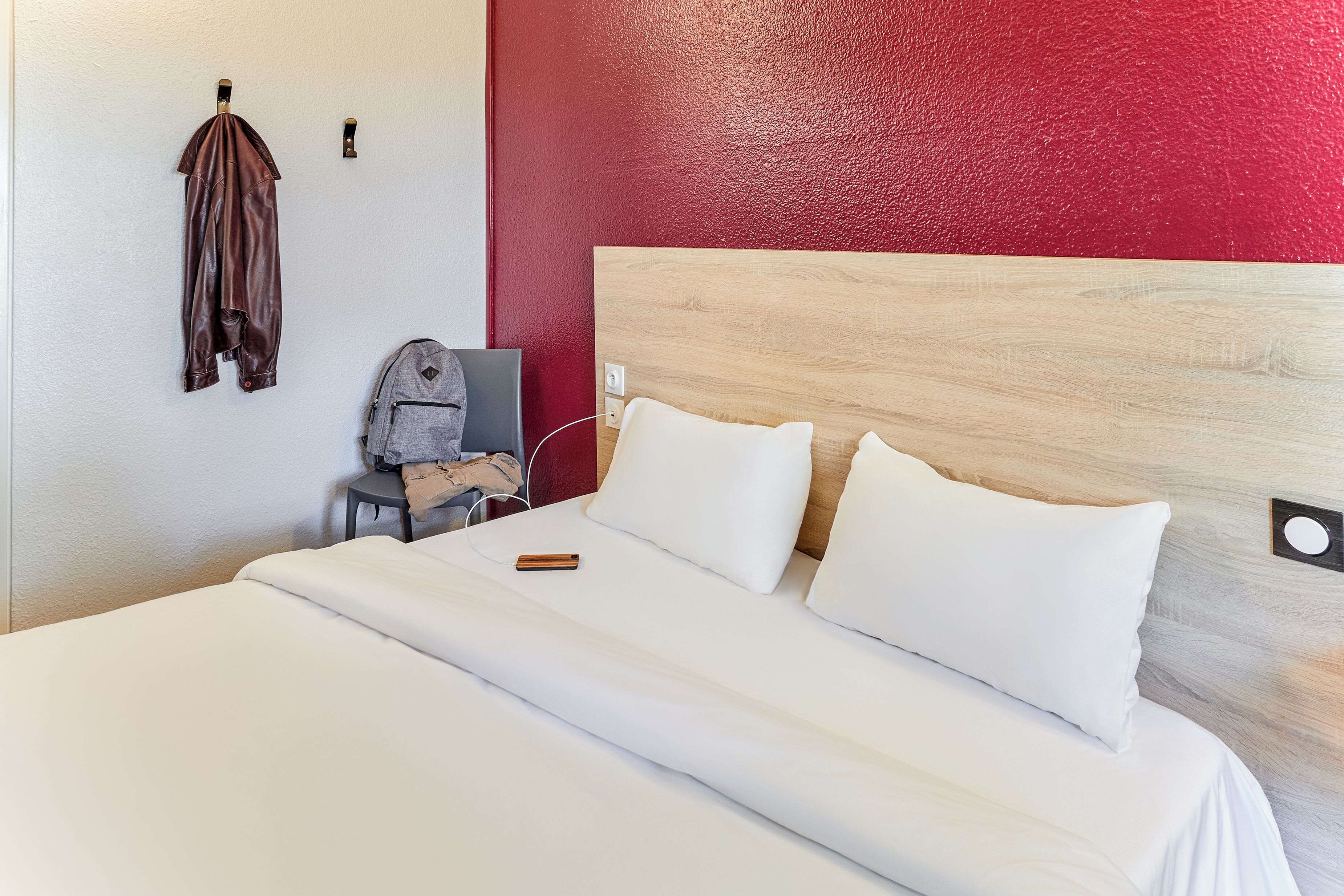 A hotel room with a bed, white bedding, a red accent wall, a wooden headboard, and a coat hanging on the wall.