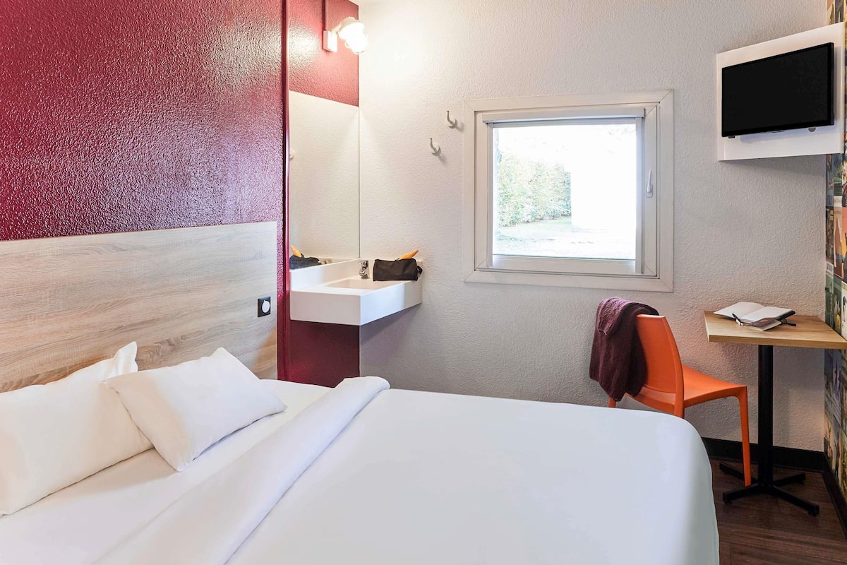Ett hotellrum med en säng, ett skrivbord med en stol, ett handfat och en tv som är monterad på väggen.