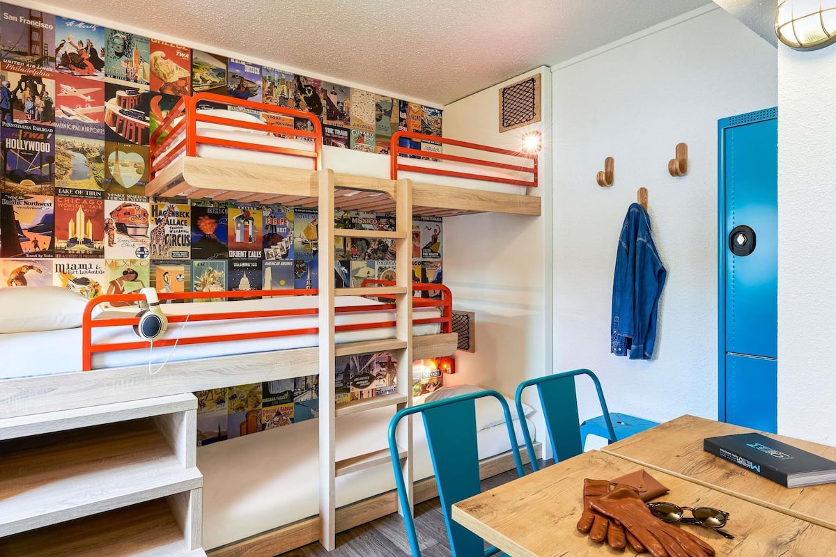 Una cama individual superpuesta con un mural con motivos de viaje, un escritorio, una silla y una mesa con un libro y guantes.