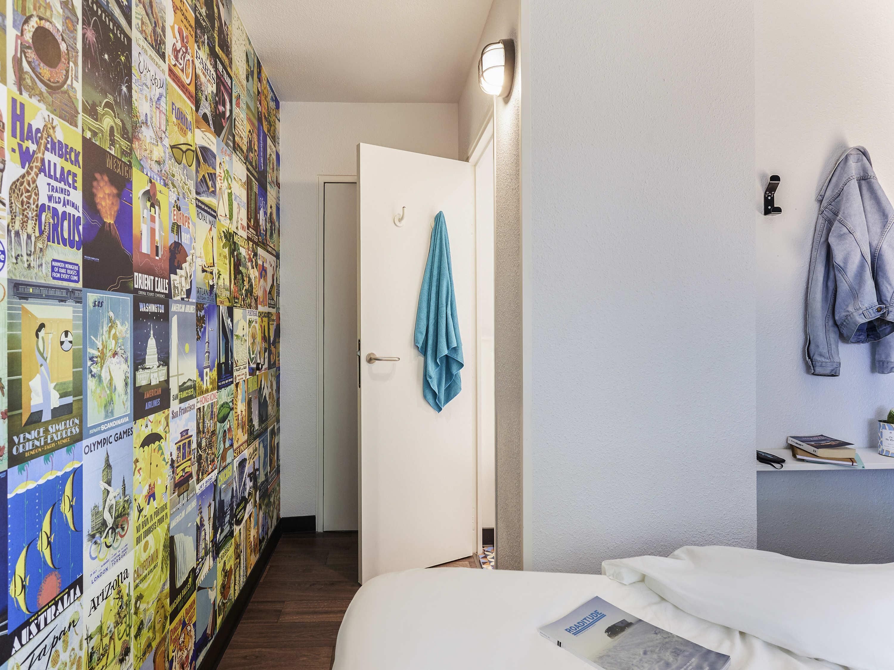 A hotel room with a wall of travel posters, a bed with a magazine, a door with a towel, and a light fixture.