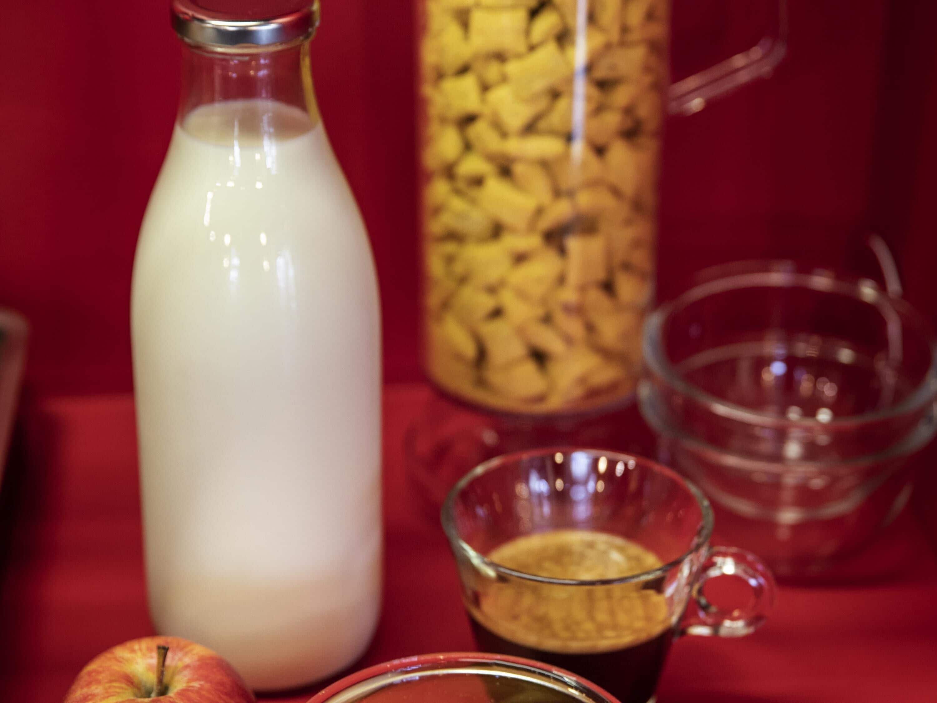A bottle of milk, a glass mug with a beverage, a glass bowl, and an apple on a red surface.