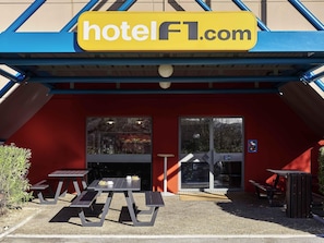A hotel entrance with a red exterior, a yellow sign reading 'hotelFL.com', and a covered outdoor seating area with tables and benches. - hotelF1 Mulhouse Bâle Aéroport (Saint-Louis)