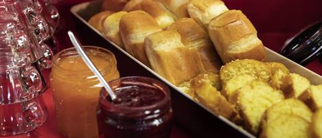 A tray of bread rolls and pastries, a jar of jam, and a glass pitcher on a red table.