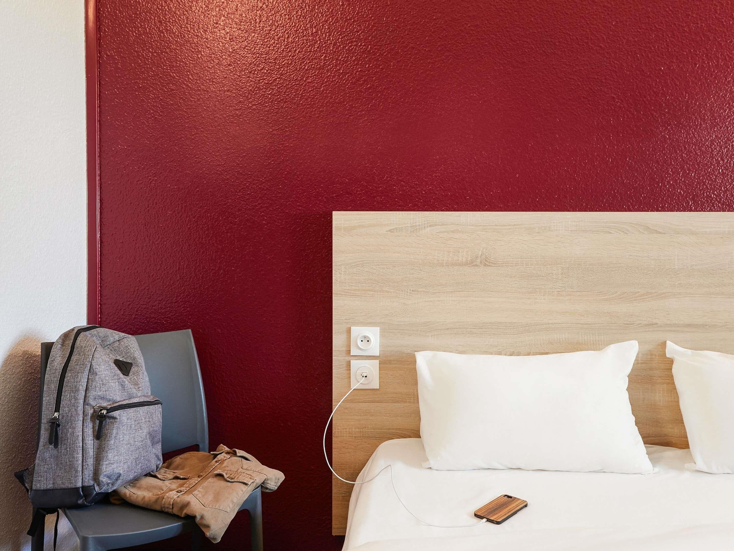Ein Hotelzimmer mit einer roten, strukturierten Wand, einem Holzkopfteil, einem Bett mit weißen Kissen, einem grauen Rucksack auf einem Stuhl und einem Smartphone auf dem Bett.