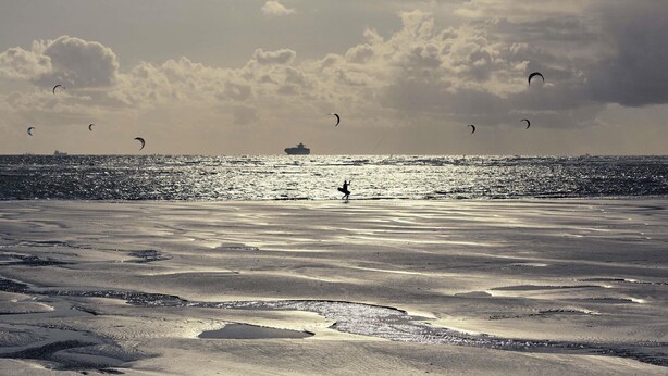 A beach with sand and water, a ship in the distance, and several kites in the sky.