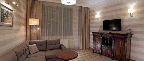 A living room with a sectional sofa, a round wooden coffee table, a flat-screen TV, a cabinet, and a large window with curtains.
