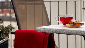 Un balcón con una mesa y una silla, un paño rojo y una taza de vidrio con una bebida roja. Al fondo, un edificio con una aguja verde.