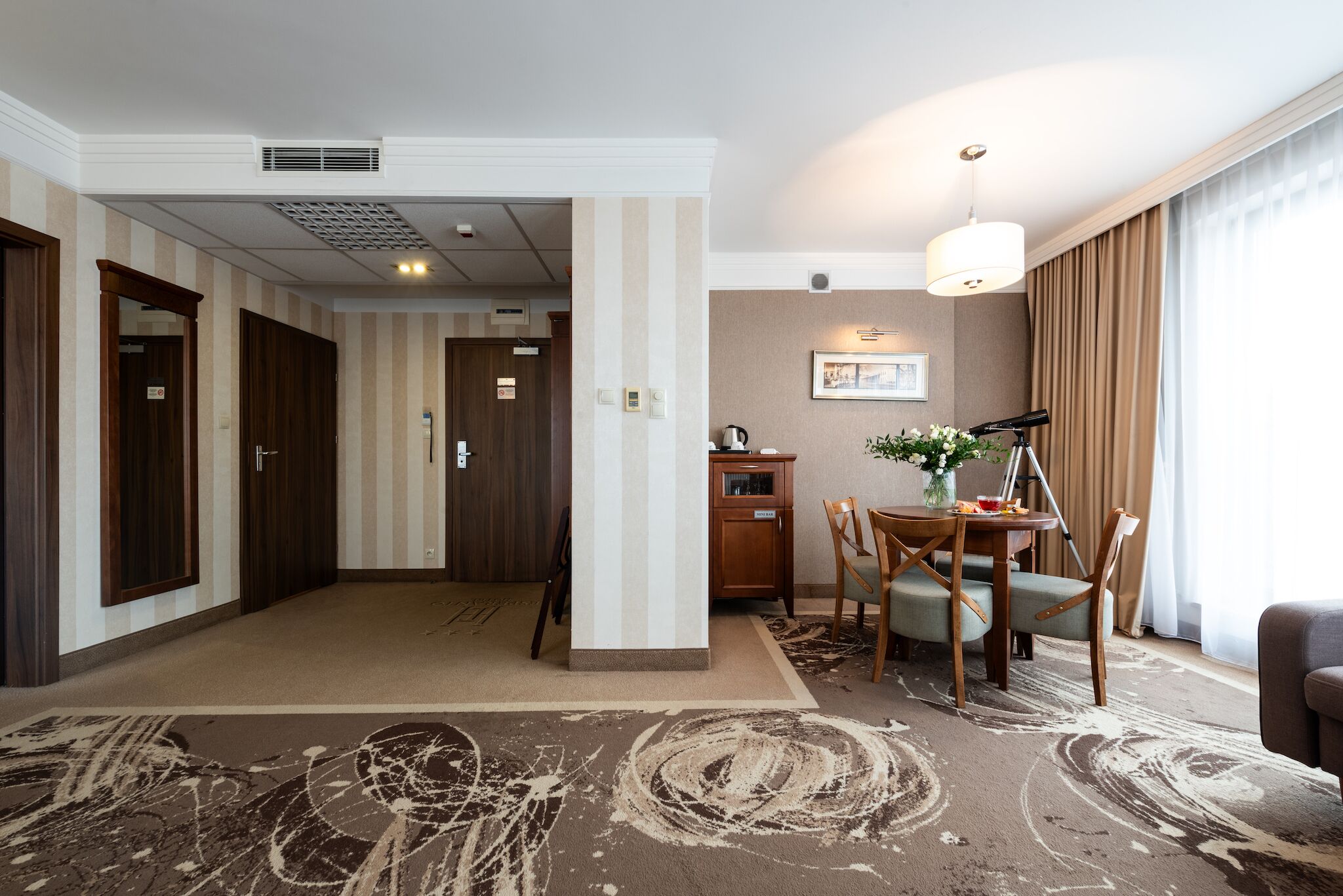 A hotel room with a dining area, a large rug, and a view of the hallway.