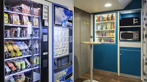 A kitchenette with a microwave, shelves stocked with snacks, and a vending machine with beverages and snacks. - hotelF1 Cholet (Cholet)