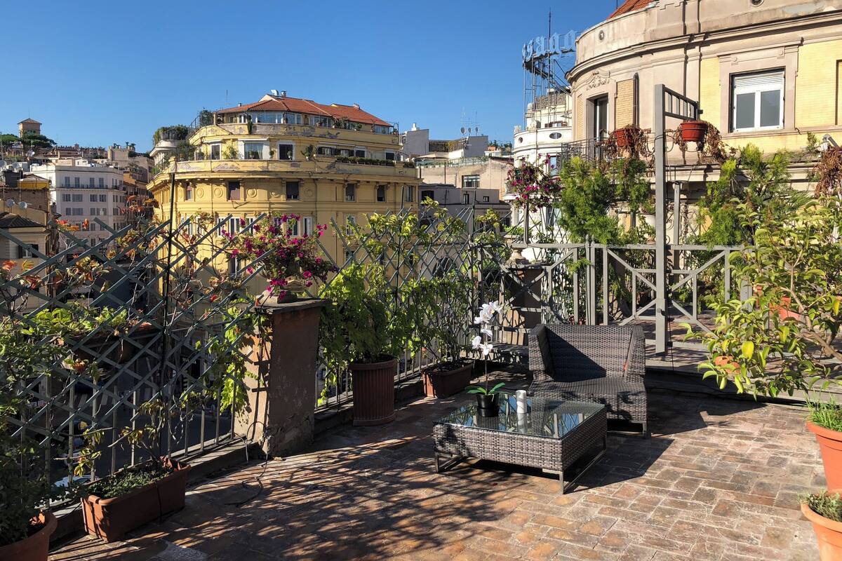 A rooftop terrace with potted plants, a glass table, and outdoor furniture.