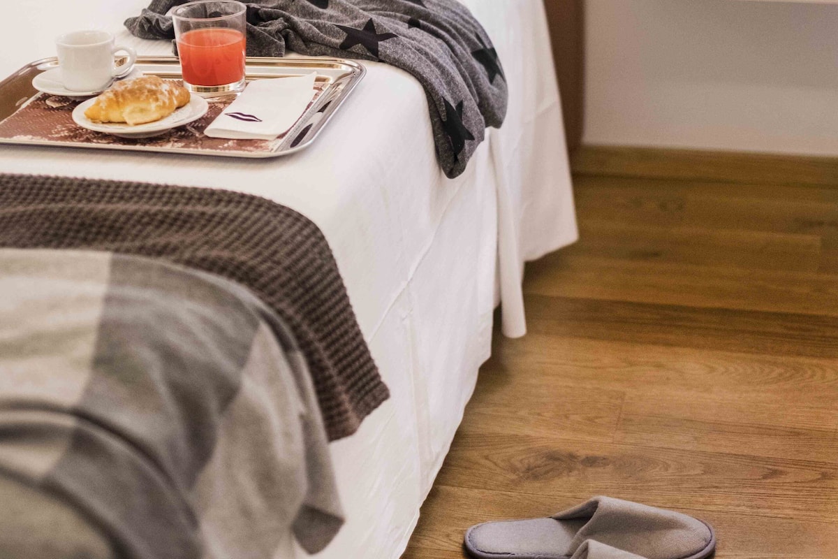 A neatly made bed with a tray of breakfast items, a bedside table with a lamp, and slippers on the floor.