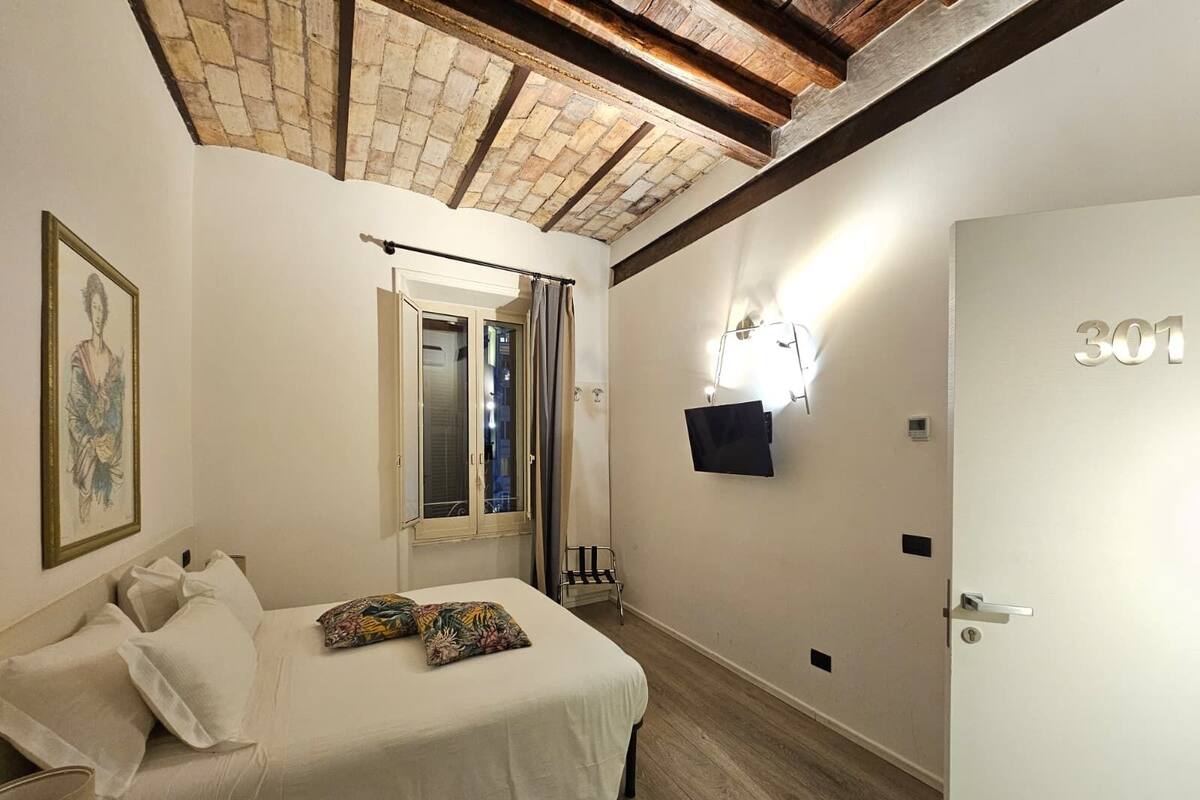 A bedroom with a bed, a window with curtains, a wall-mounted light, and a door with a number.