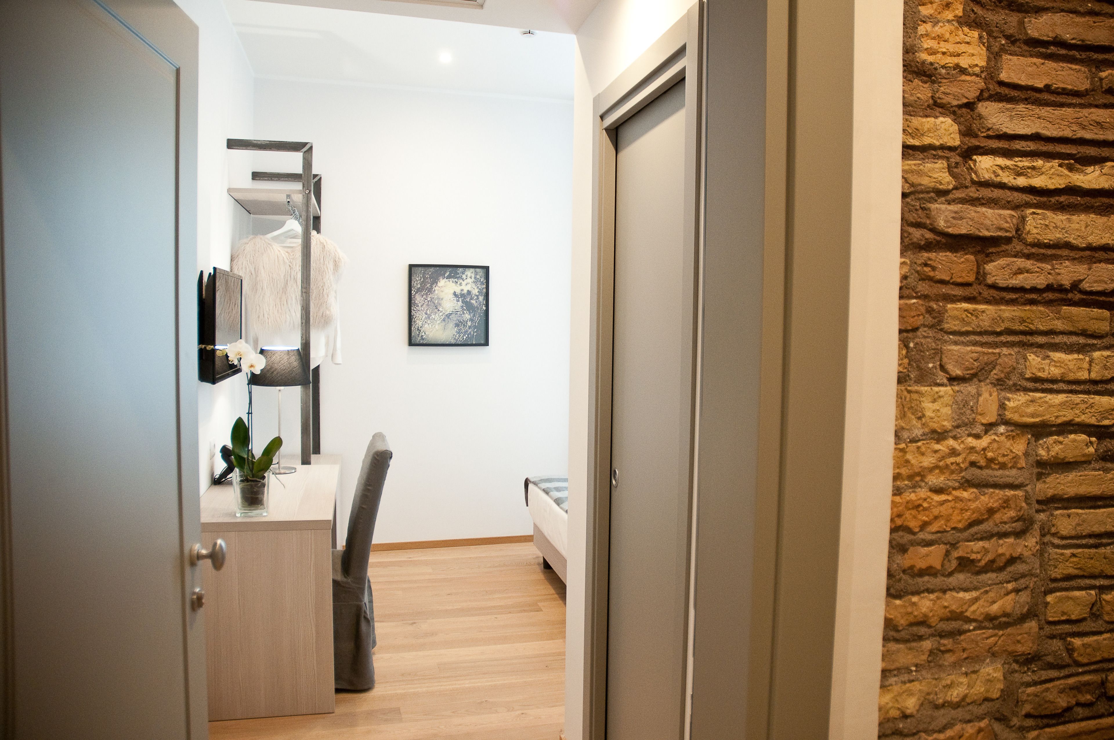 Een kamer met een bed, een bureau met een plant, een stoel, een garderobe en een wandplank met een jas.