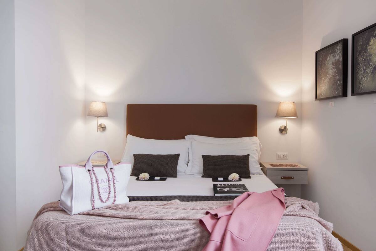 A neatly made bed with a brown headboard, two bedside lamps, and a nightstand with a book and a small table.