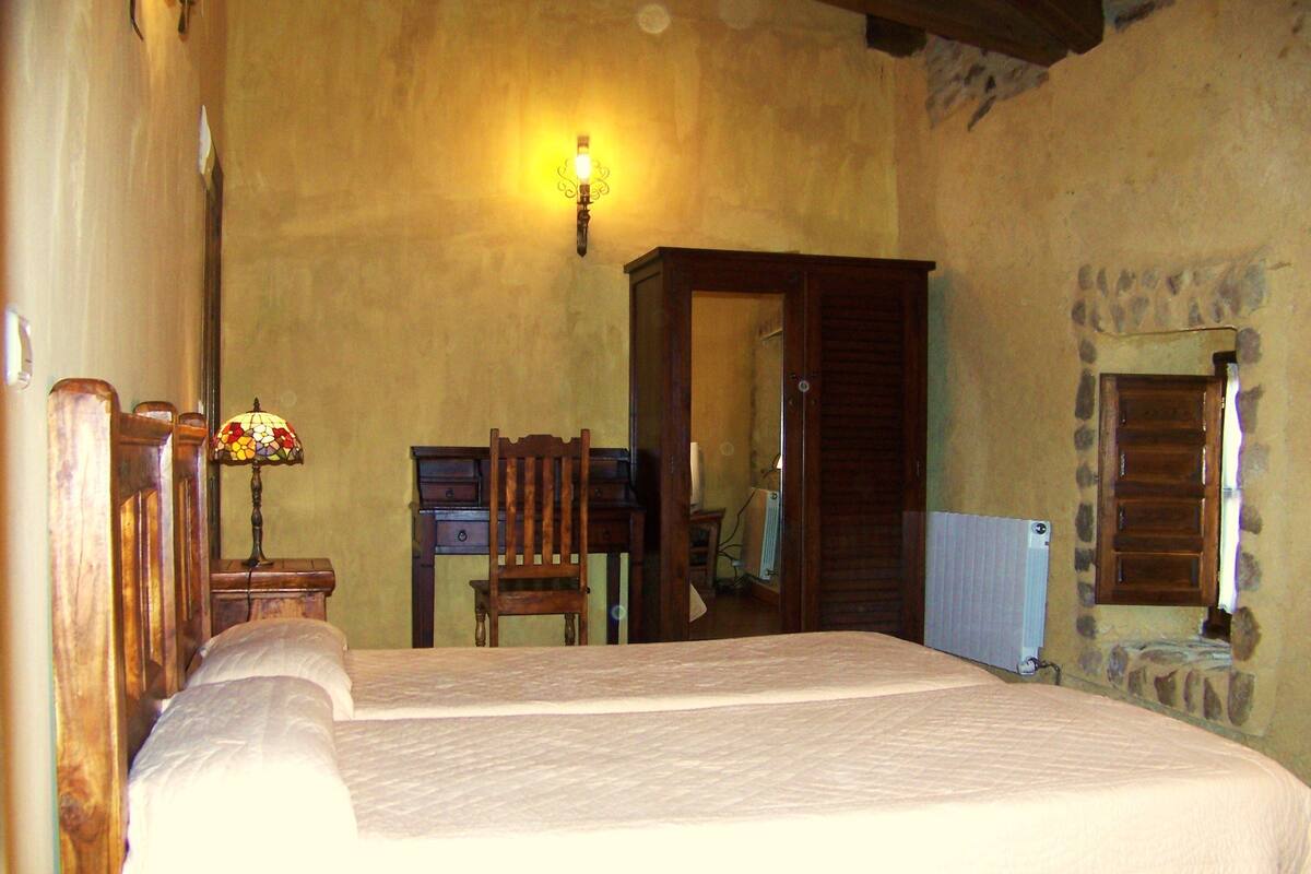 A bedroom with a large bed, a wooden desk, a chair, a lamp, and a stone wall.