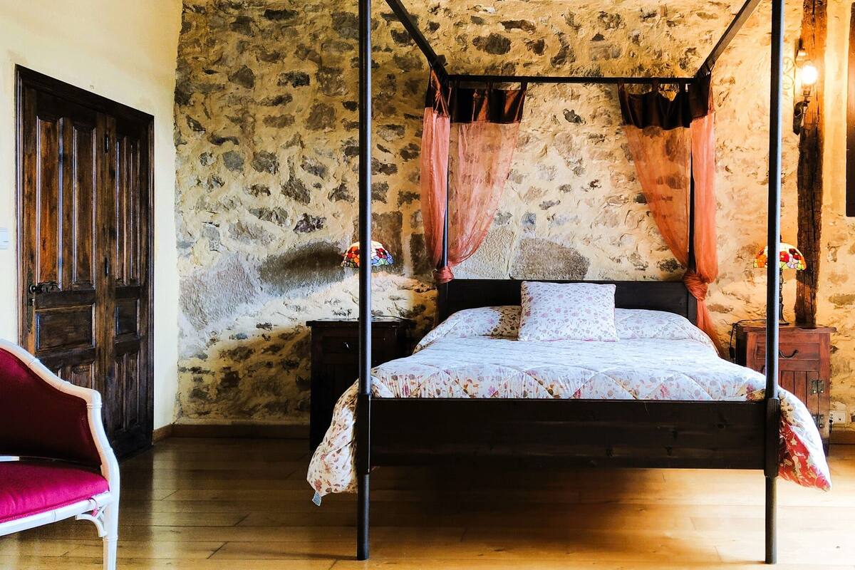 A four-poster canopy bed with a floral bedspread, a stone wall, a wooden door, and a red armchair.